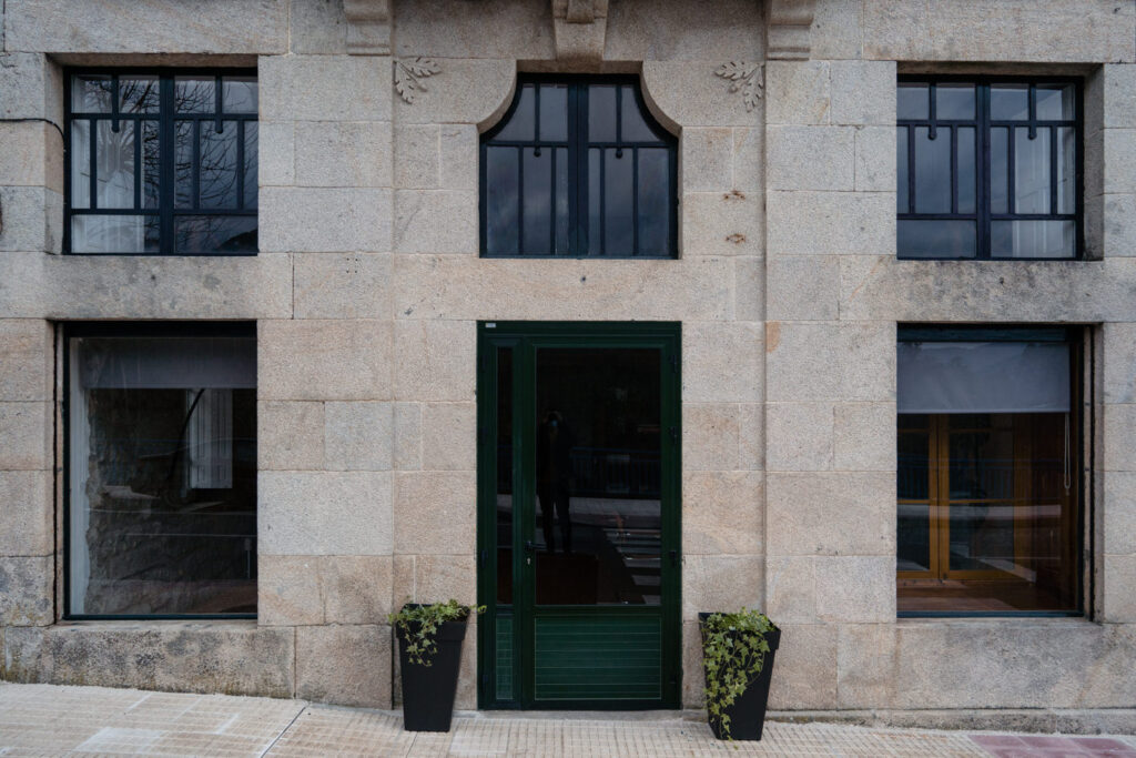 Alzado de la antigua escuela: fachada de sillería de granito con carpinterías negras y puerta verde.